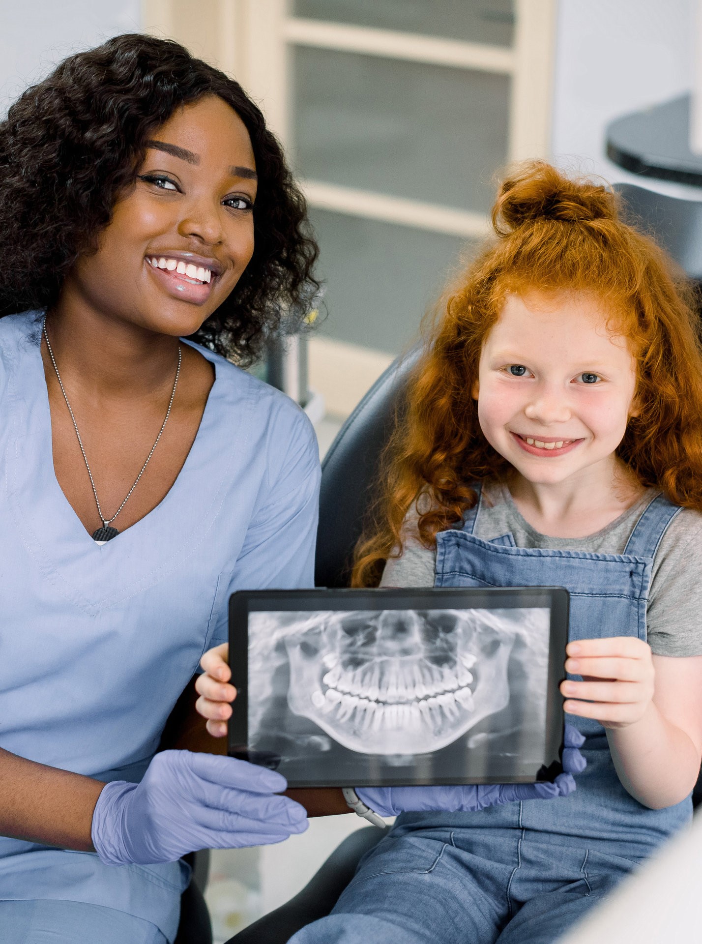 Little smiling girl with red curly hair sitting on chair and looking at camera, while holding x-ray scan image of her teeth on digital tablet together with her cheerful black female dentist at clinic