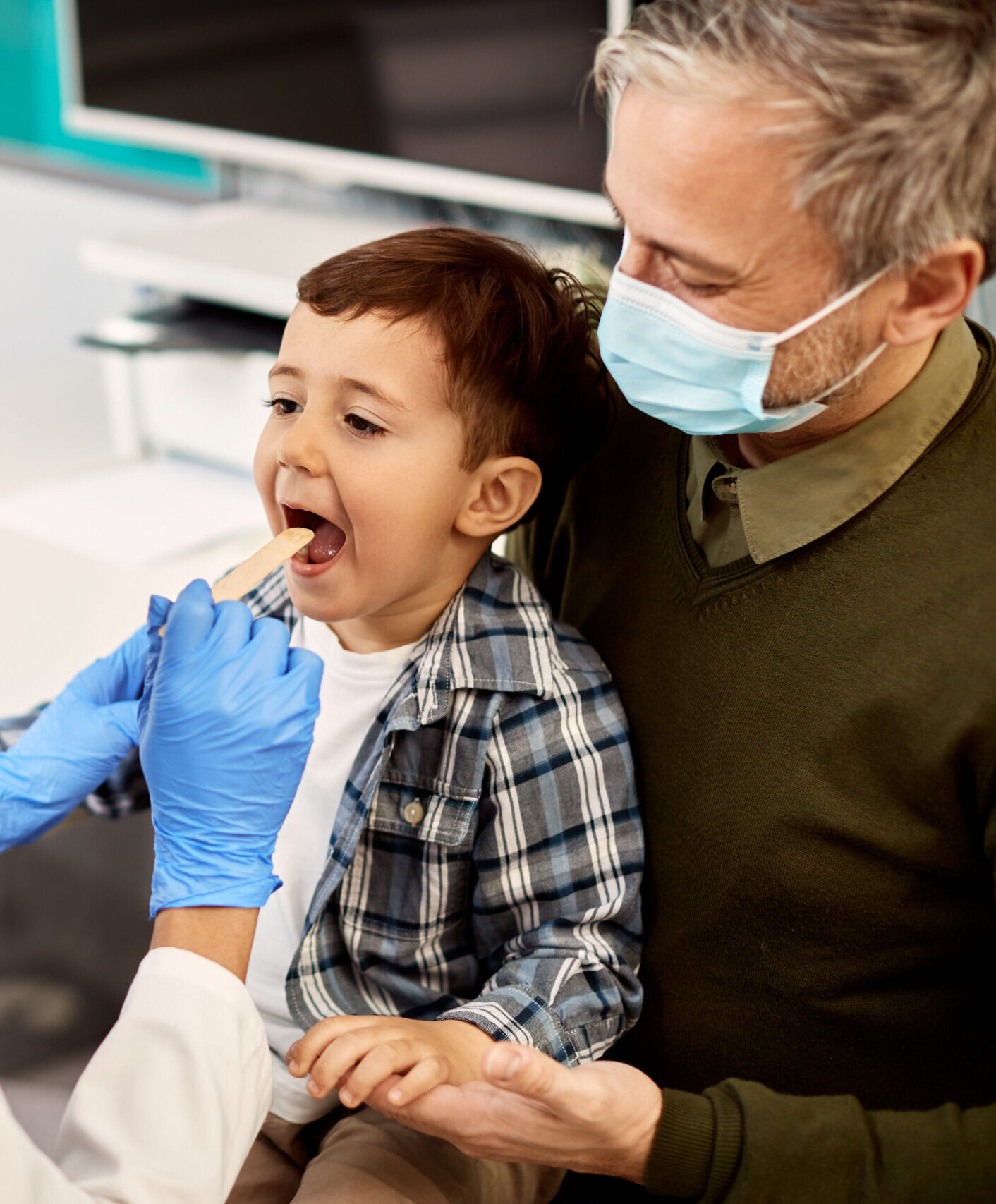 African American female dentist examining teeth of a small boy who is sitting on father's lap during dental appointment. Focus is on boy.