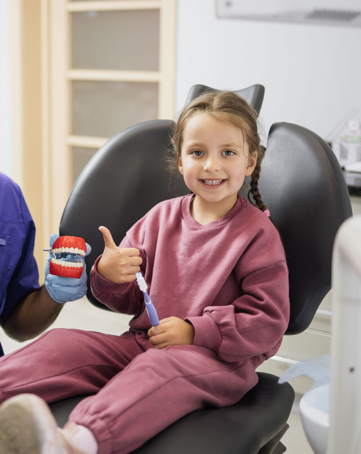 Happy confident male dentist tells little child girl how to brush the teeth on artificial jaw model showing thumbs up. Caries prevention, pediatric dentistry, milk teeth hygiene concept.