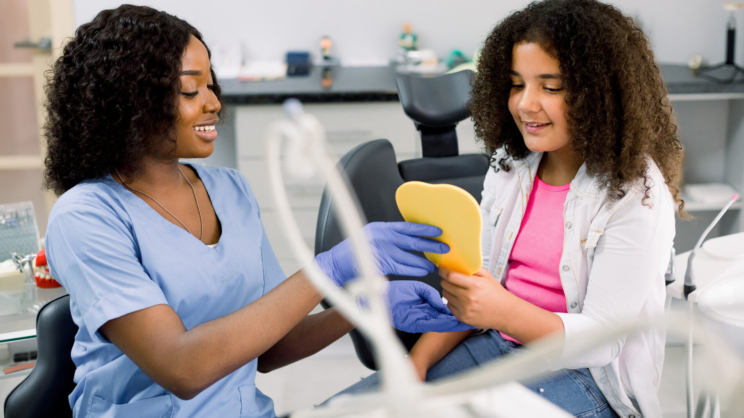 Cute happy mixed raced teen girl, looking at the mirror with toothy smile, sitting on the dental chair while smiling black female dentist showing the result of treatment teeth at the dental office.