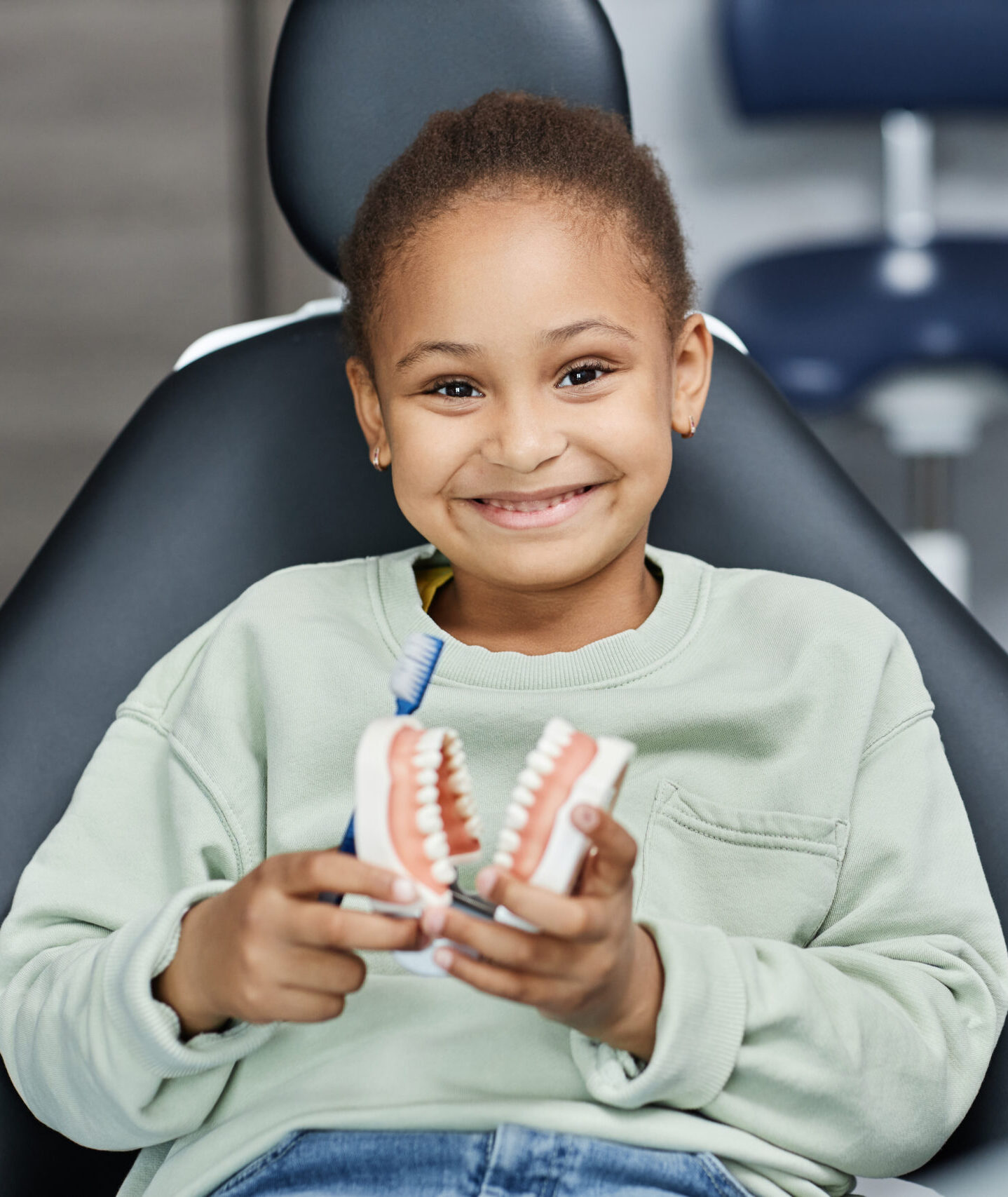 Front view portrait of black little girl in dental chair smiling at camera happily and holding tooth model, copy space