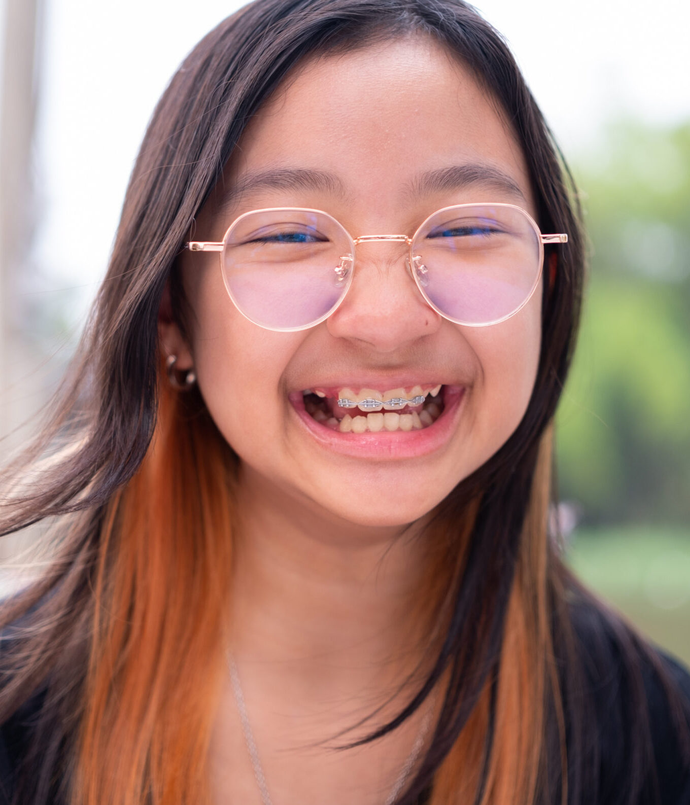Asian girl teenage girl with braces and eyeglasses wearing black t-shirt smiling brightly on background outdoors.