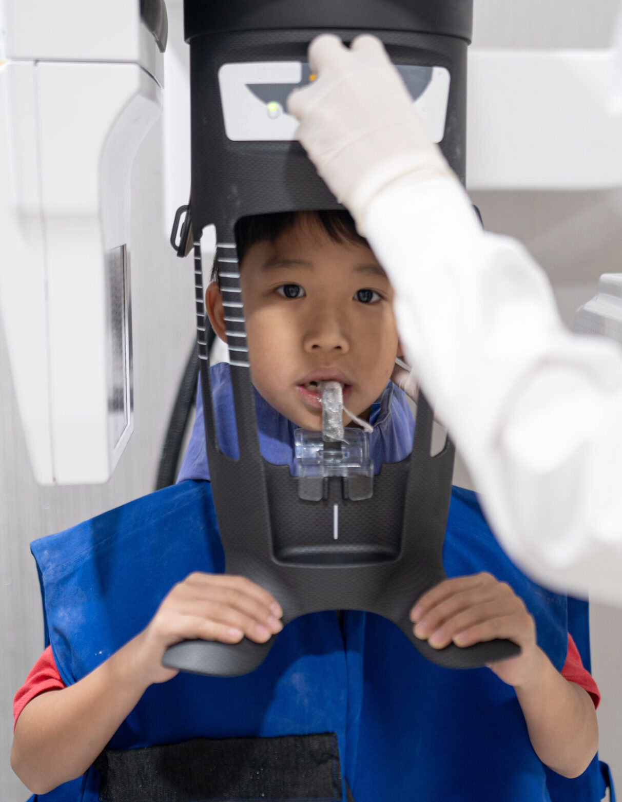 Dental surgery X-ray examination, patient at stomatology clinic. A young boy wearing blue safety gear sits in front of dental equipment while an x-ray is taken.